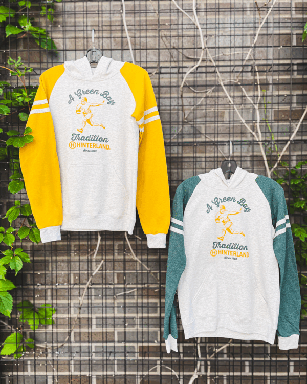 Two hoodies hanging on a fence. One has yellow sleeve and one has green sleeves. They both have a retro football player on them and the phrase "A Green Bay Tradition."