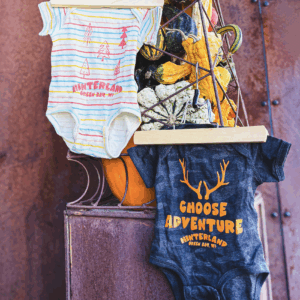 Two baby onesies on hangers with Hinterland branding. One is rainbow striped and the other is camo.